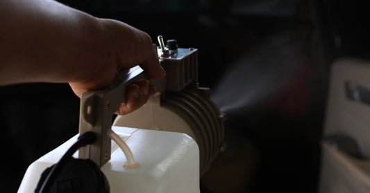Close-up of a hand carrying a fogging machine with a white plastic tank while spraying chemicals inside a dark room.