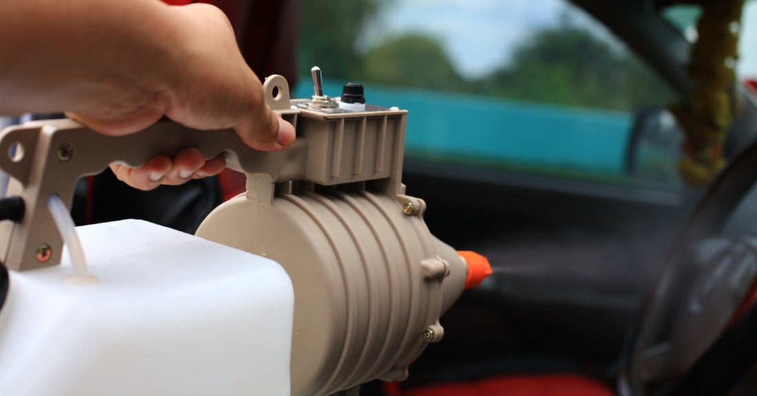 A hand holding a chemical fogger attached to a white tank and a brown atomizer, spraying a fine mist into a car.