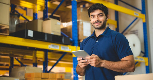 Young man wearing a blue polo, holding a tablet with both hands standing inside a large warehouse with boxes.