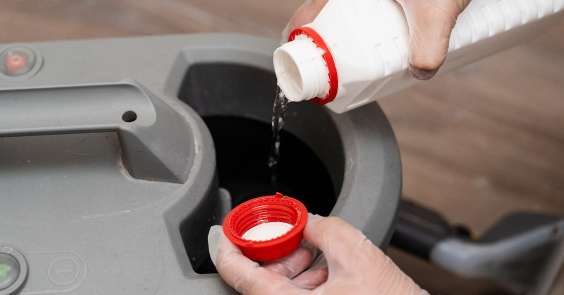 Close-up of a hand holding a red lid pouring chemicals from a white bottle into a grey chemical fogger placed on the ground.